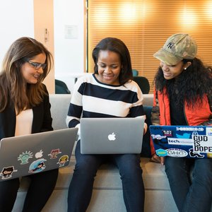 3 women with laptops balanced on their knees are pointing at their screens and talking and smiling