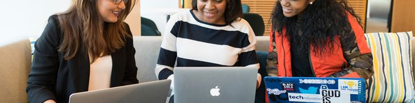3 women with laptops balanced on their knees are pointing at their screens and talking and smiling