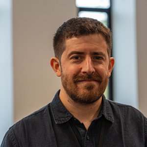 A head and shoulders photo of Martin.  A male with short brown hair and a short full beard wearing a collared shirt standing in a white walled office with vertical windows.