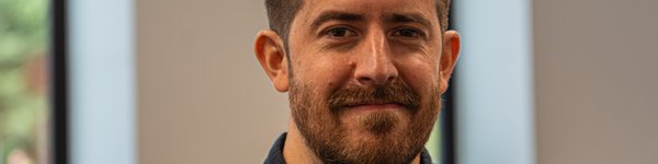 A head and shoulders photo of Martin.  A male with short brown hair and a short full beard wearing a collared shirt standing in a white walled office with vertical windows.
