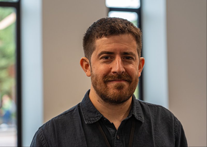 A head and shoulders photo of Martin.  A male with short brown hair and a short full beard wearing a collared shirt standing in a white walled office with vertical windows.